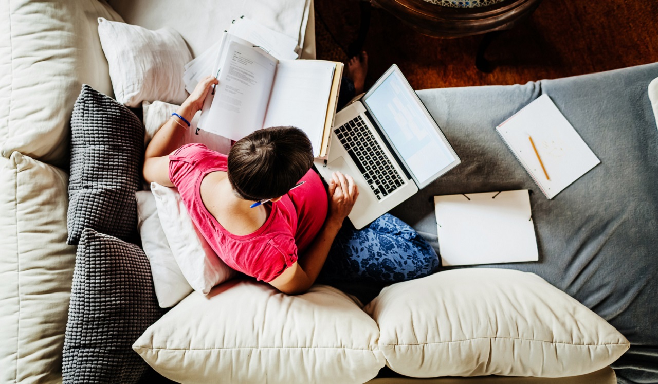 A woman on the couch with a book, notebook and laptop computer.