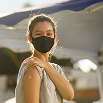 A teenage girl wearing a face mask shows the band-aid from her vaccine shot.