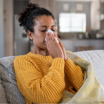 A woman holds a tissue to her nose as she sits on the couch.