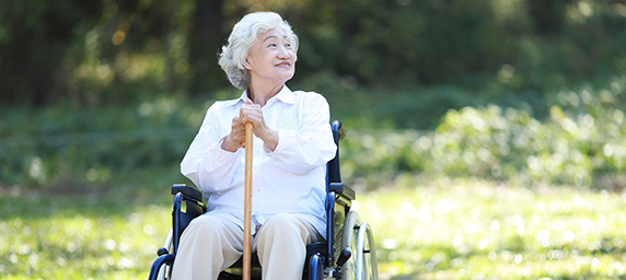 A woman in a wheel chair holds a cane while sitting in the sun, looking up hopefully.