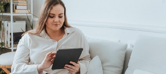 A woman sits on her couch with an electronic tablet in her hands. 